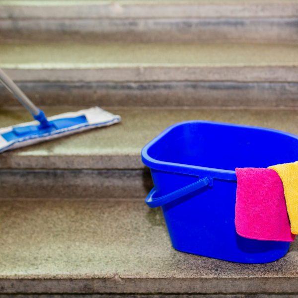 A blue plastic bucket with yellow and pink microfiber cloth, standing on grey granite stairs. A hand holding mop wiping the stairs.
